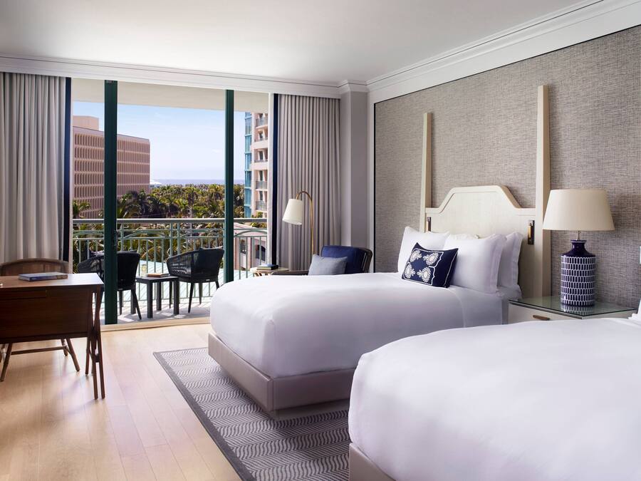 Habitación de hotel con dos camas, un escritorio y un balcón con vistas a edificios y palmeras.