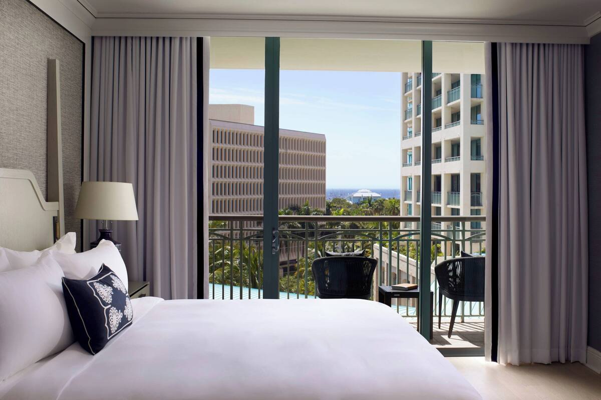 A hotel room with a large bed, a nightstand with a lamp, and a view of a balcony with chairs and a building in the background.