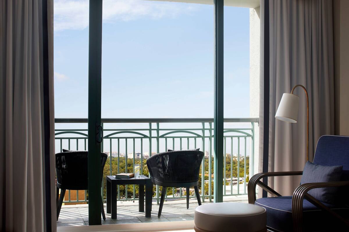 A balcony with a view of the city, furnished with a chair, a small table, and a lamp.