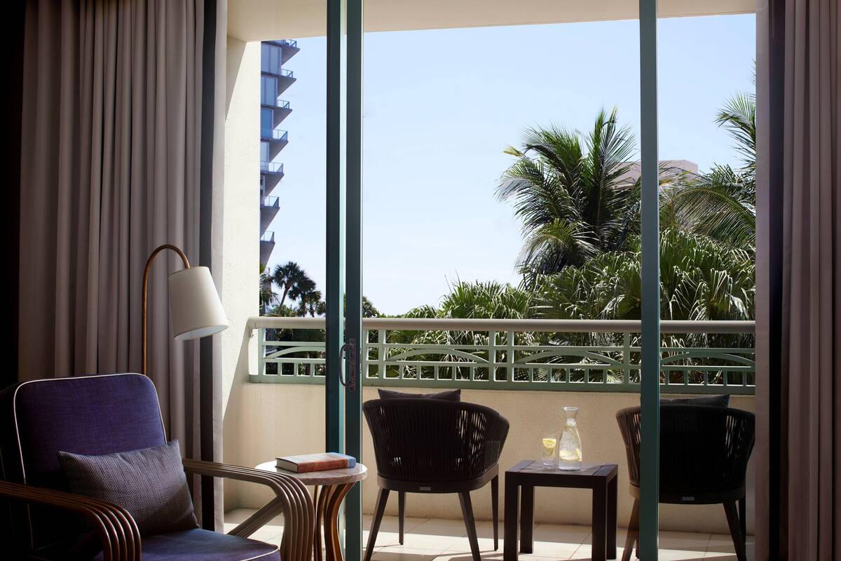 A balcony with a view of palm trees and a building. There are two chairs, a table, and a lamp.