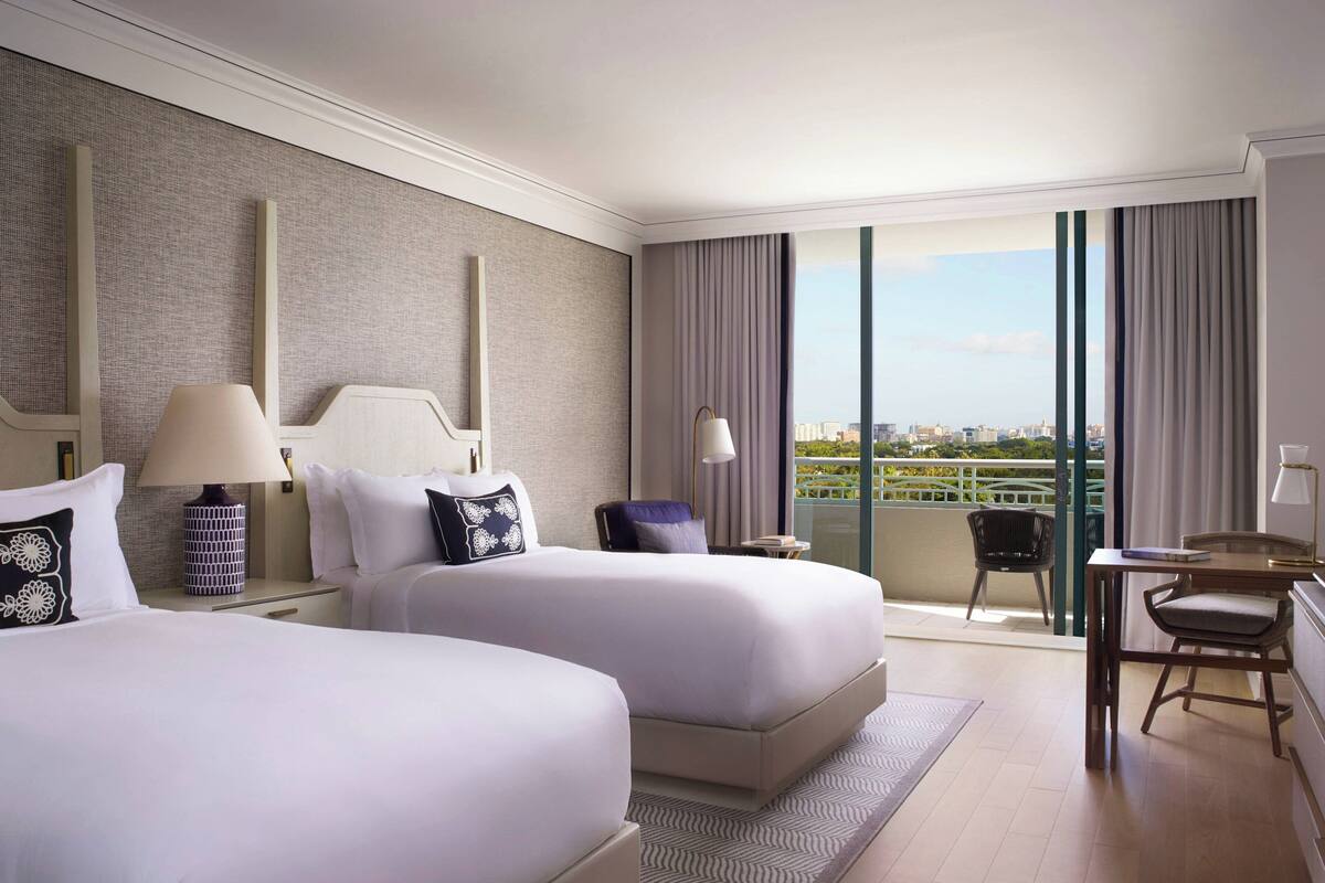 A modern hotel room with two beds, a desk, and a balcony with a city view.