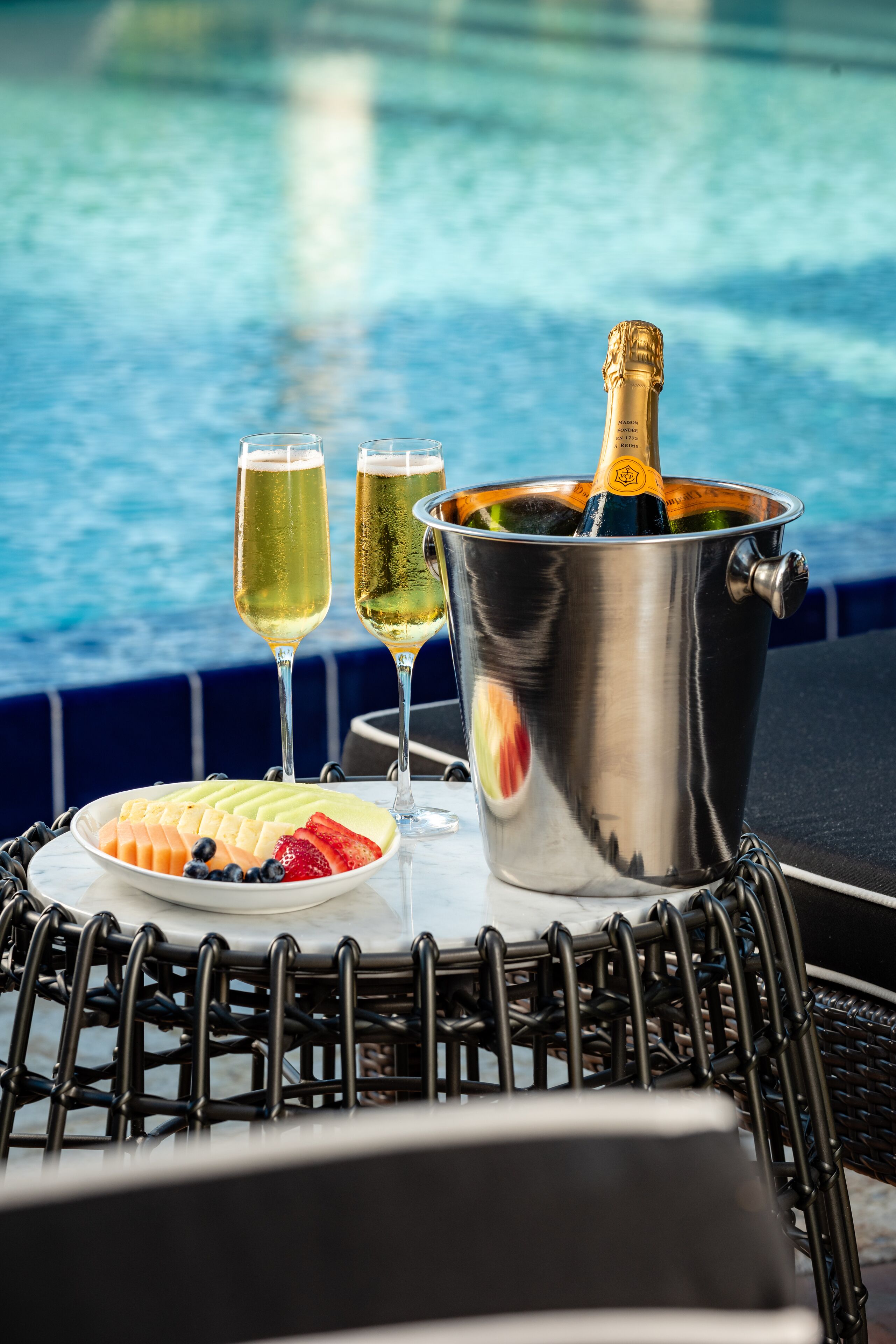 A metal table with a bottle of champagne, two glasses, and a plate of fruit by a poolside.