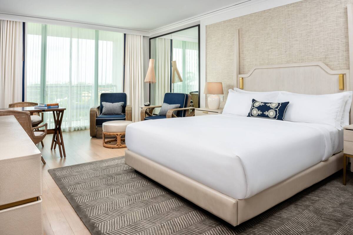 A hotel room with a large bed, two armchairs, a desk, and a view of the city through large windows.