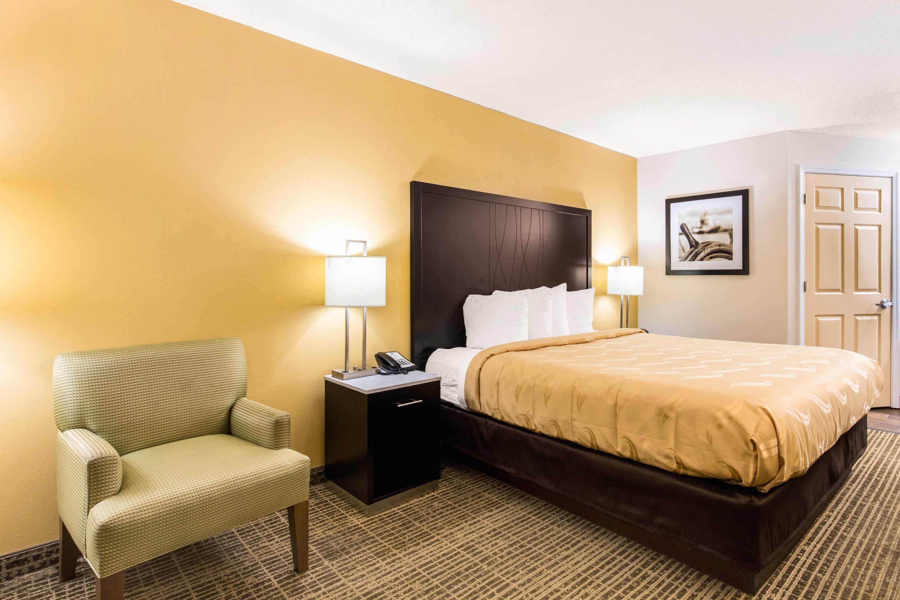 Una habitación de hotel con una cama, una silla, una mesita de noche con lámpara y una puerta.
