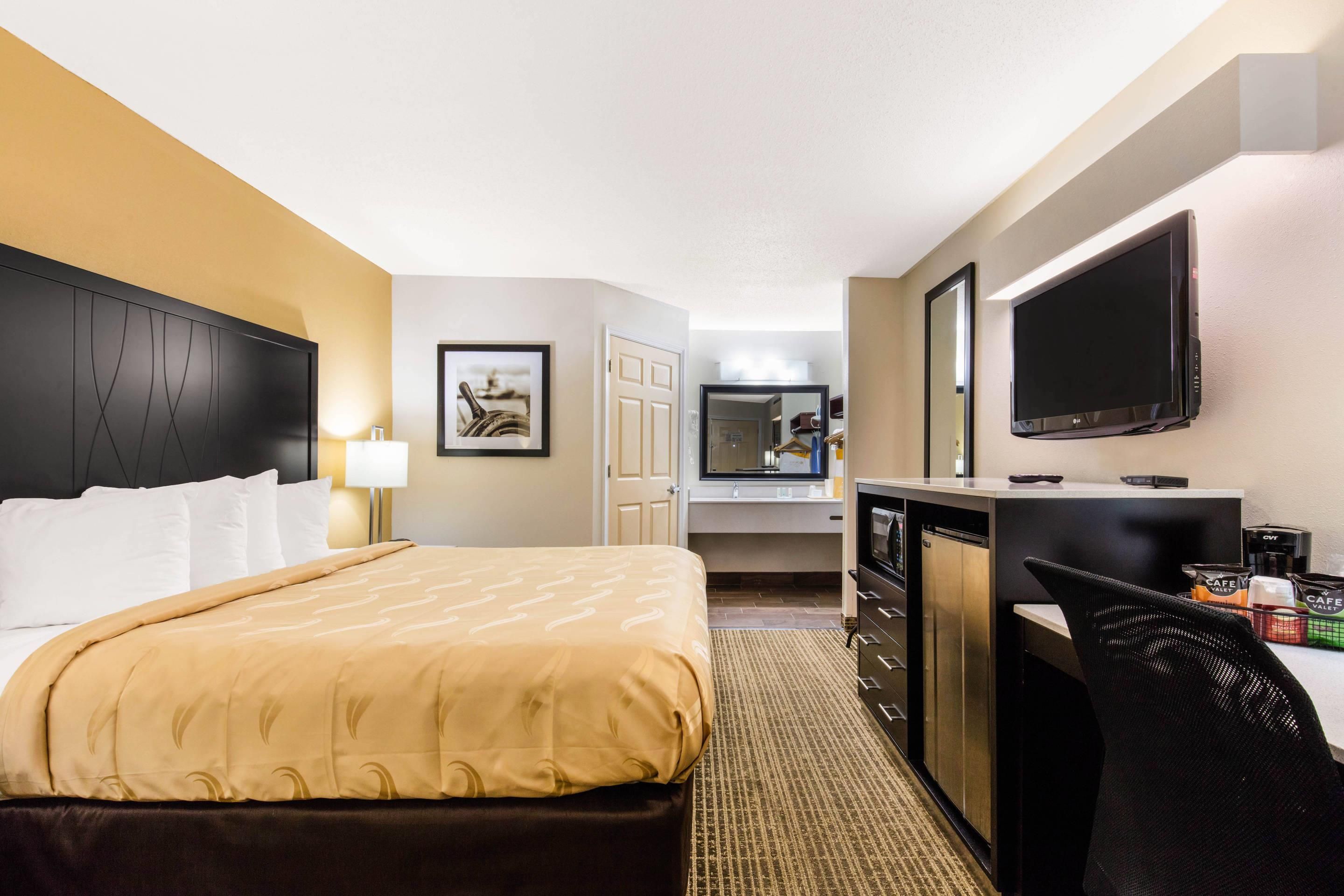 A hotel room with a bed, a desk with a flat-screen TV, a coffee machine, and a mirror.