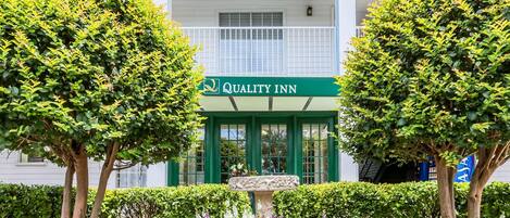 A hotel entrance with a sign reading 'Quality Inn,' surrounded by well-maintained hedges and flowering plants.