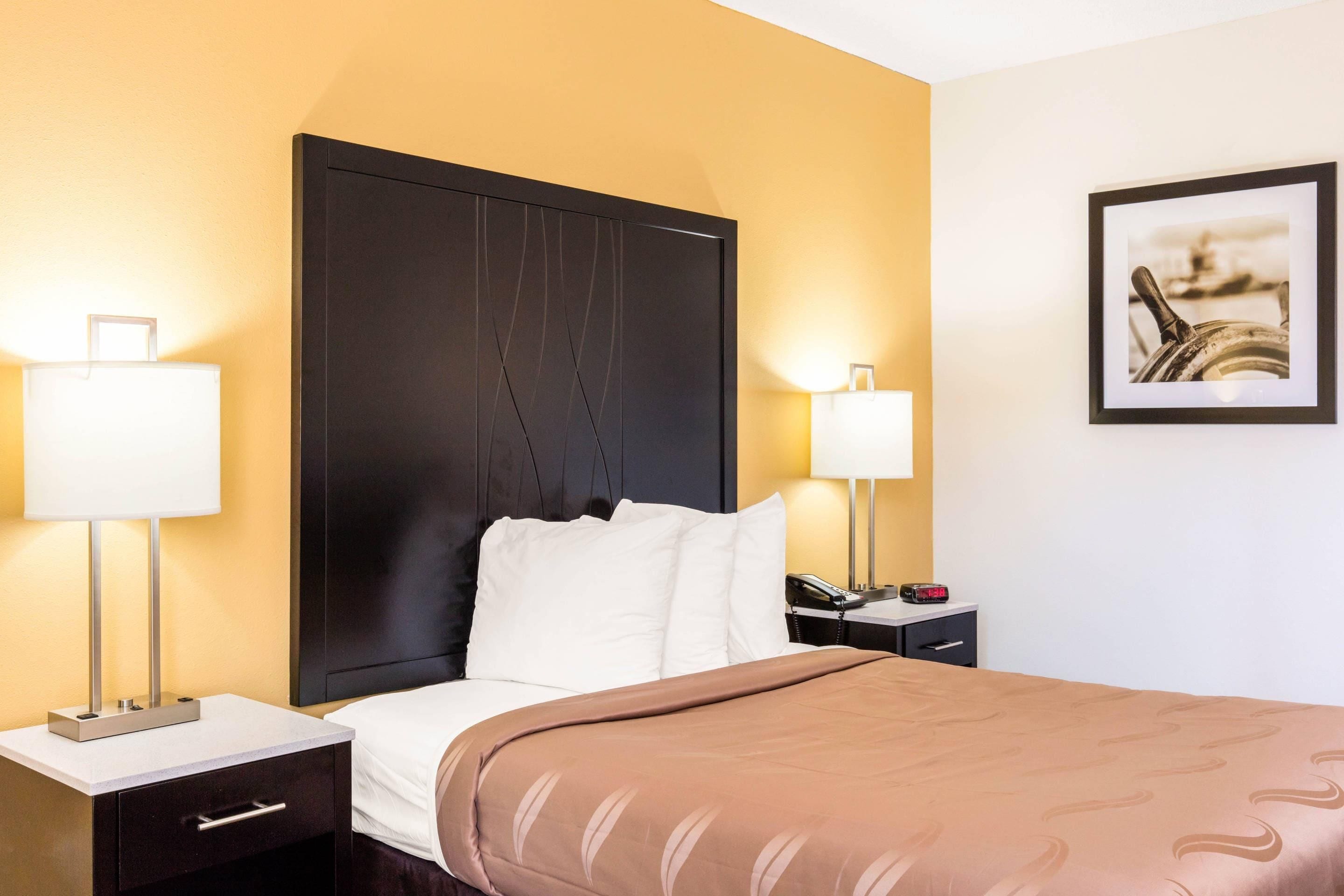 Ein Hotelzimmer mit einem großen Bett, zwei Nachttischen, einer Lampe, einer Uhr und einem gerahmten Bild an der Wand.