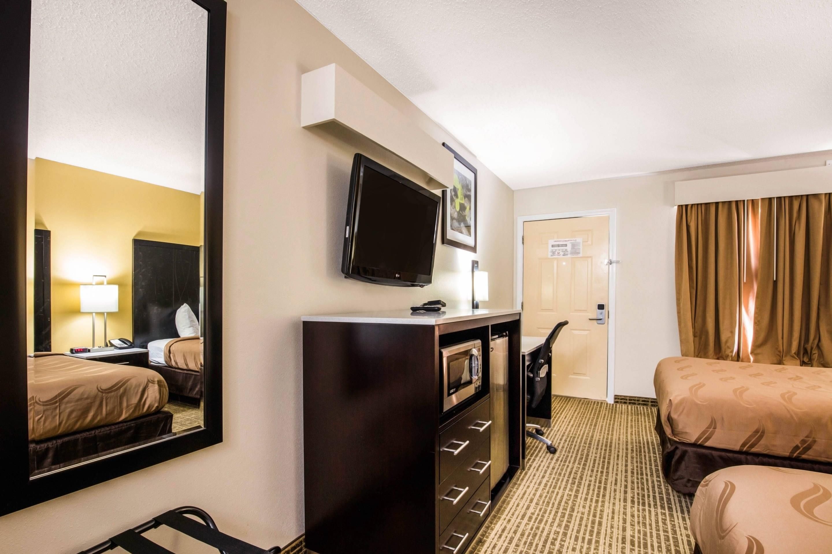 Een hotelkamer met een bed, een bureau, een televisie, een spiegel, een kast en een deur naar een andere kamer.