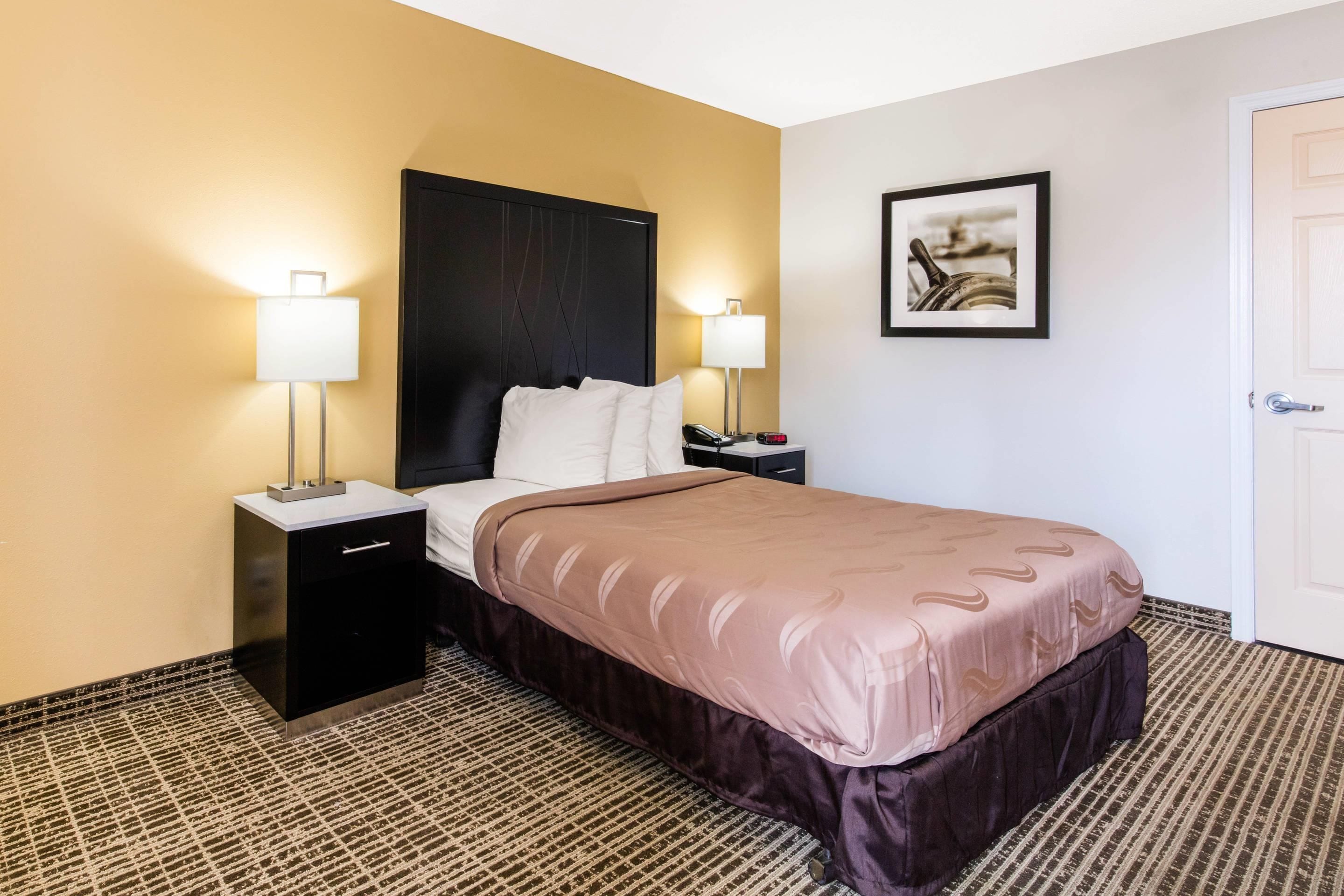 A hotel room with a bed, bedside tables, lamps, a framed picture on the wall, and a door.