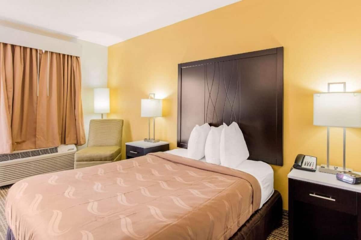 A hotel room with a bed, two bedside tables, a chair, a lamp, and a wall-mounted phone.