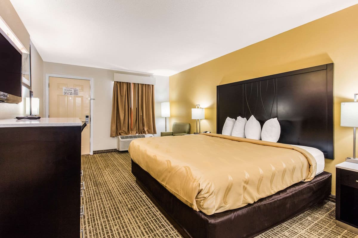 A hotel room with a large bed, a desk, and a chair. There is a door with a sign, a window with curtains, and a wall-mounted TV.