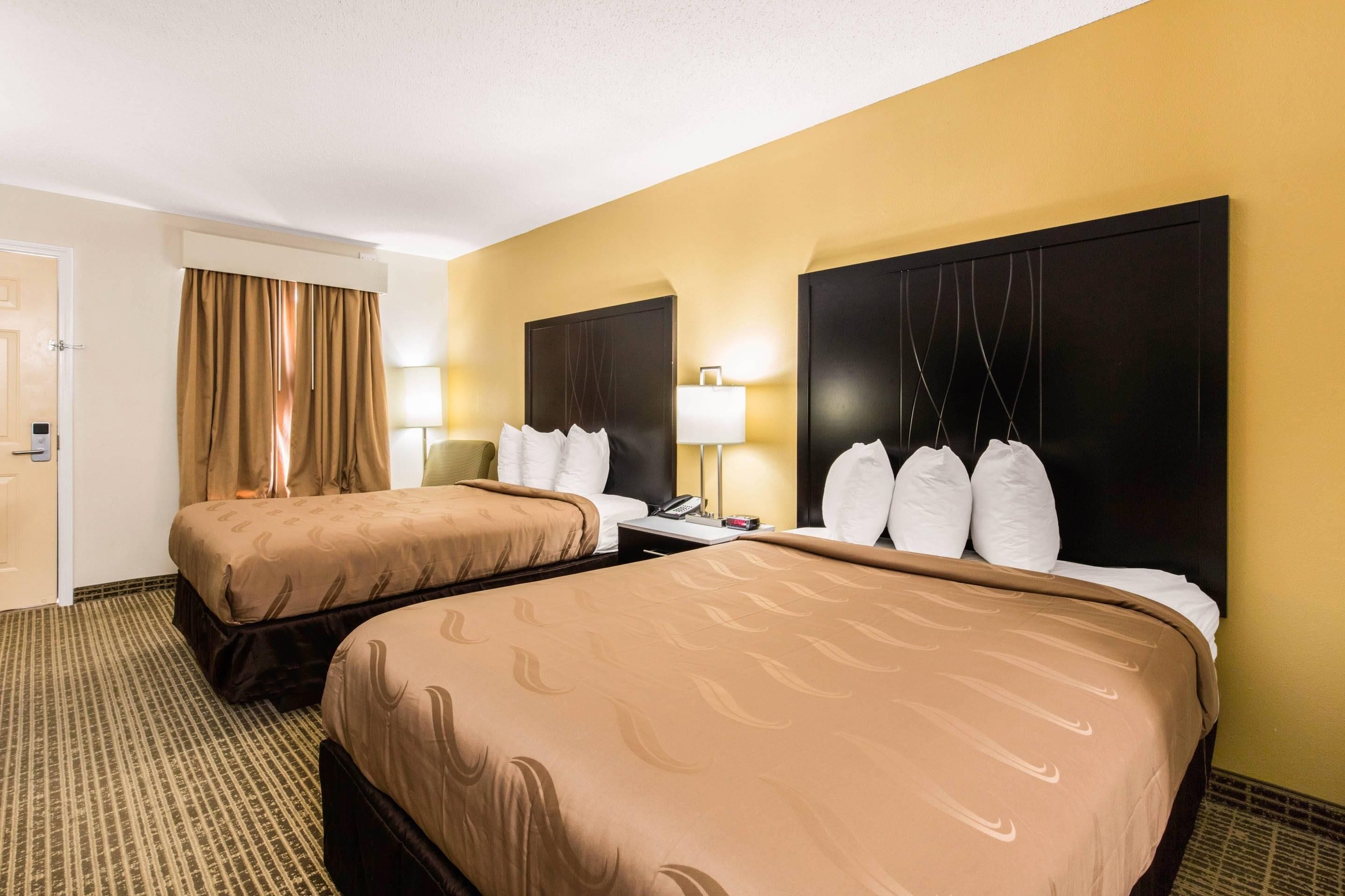 A hotel room with two beds, a door, a curtain, a nightstand with a lamp, and a wall-mounted telephone.