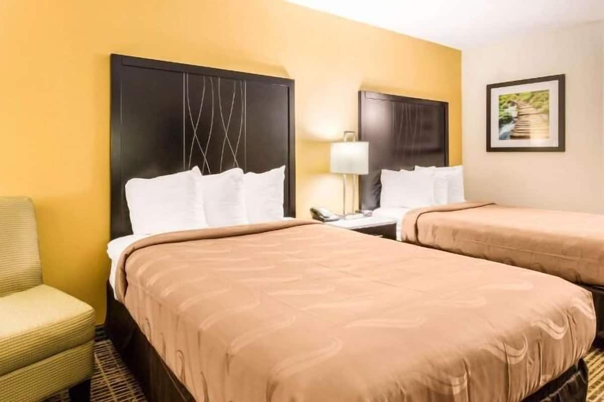 A hotel room with two beds, a chair, a nightstand, a lamp, and a framed picture on the wall.