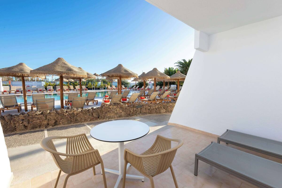 Zona junto a la piscina con sillas de descanso, una mesa redonda y sombrillas de paja.