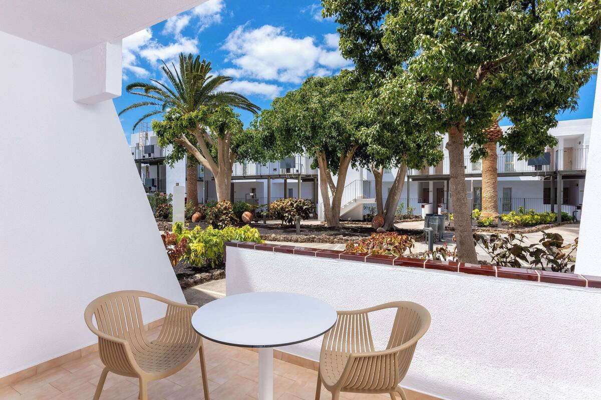 Un patio al aire libre con dos sillas de mimbre y una pequeña mesa redonda, rodeado de paredes blancas y vistas a un conjunto de edificios con árboles y plantas.