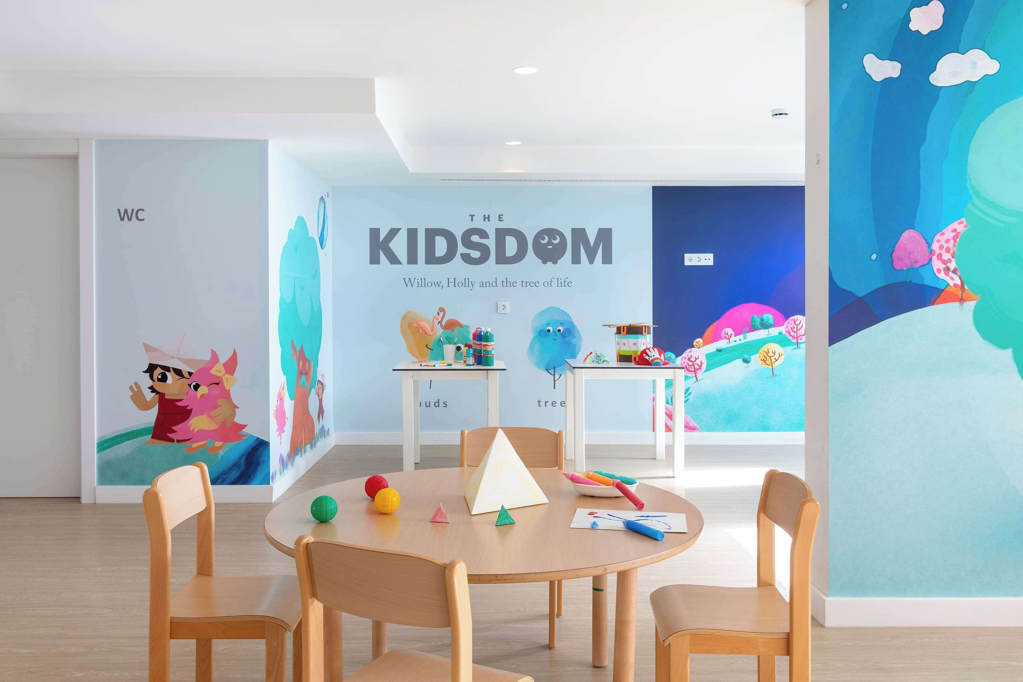 A children's playroom with a mural titled "The Kids' Room," featuring characters Willow and Holly. There are wooden chairs around a table with toys and educational materials.