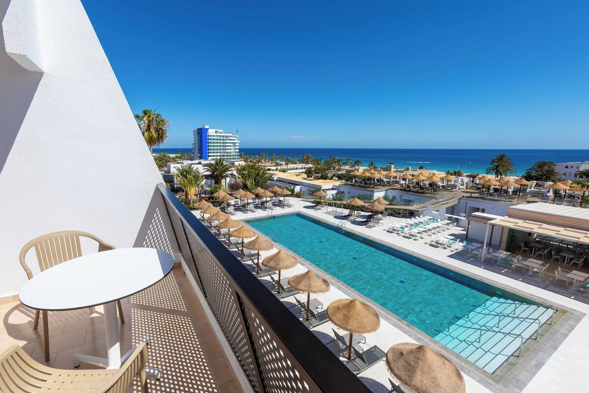 Zona junto a la piscina con sillones de descanso y sombrillas, con vista a una piscina azul cristalina y al océano.