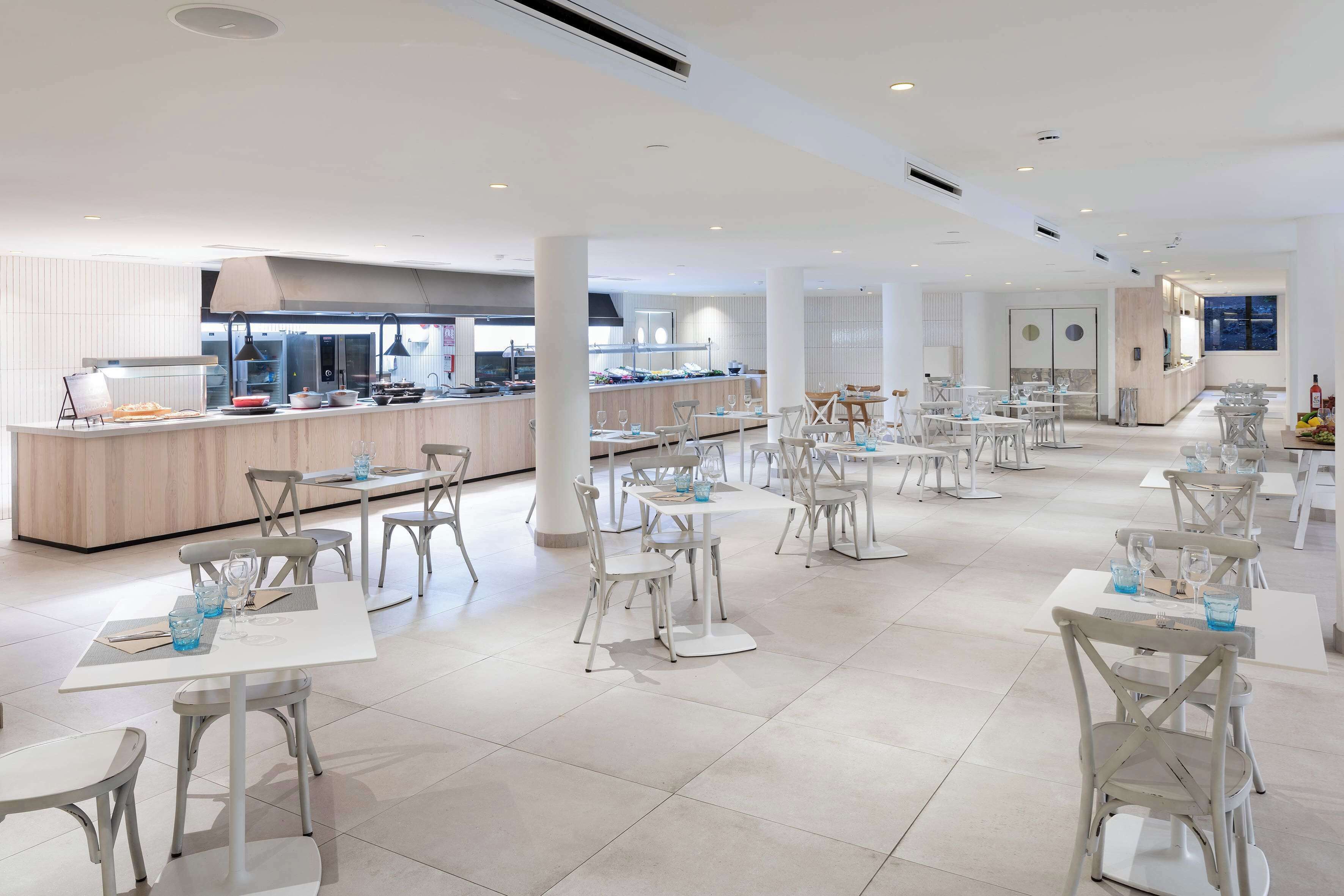 Un vaste espace de salle à manger moderne, avec des tables et des chaises blanches, un coin cuisine central et une vue sur la ville la nuit.