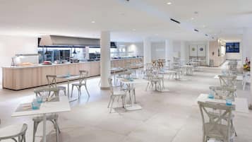 Un vaste espace de salle à manger moderne, avec des tables et des chaises blanches, un coin cuisine central et une vue sur la ville la nuit.