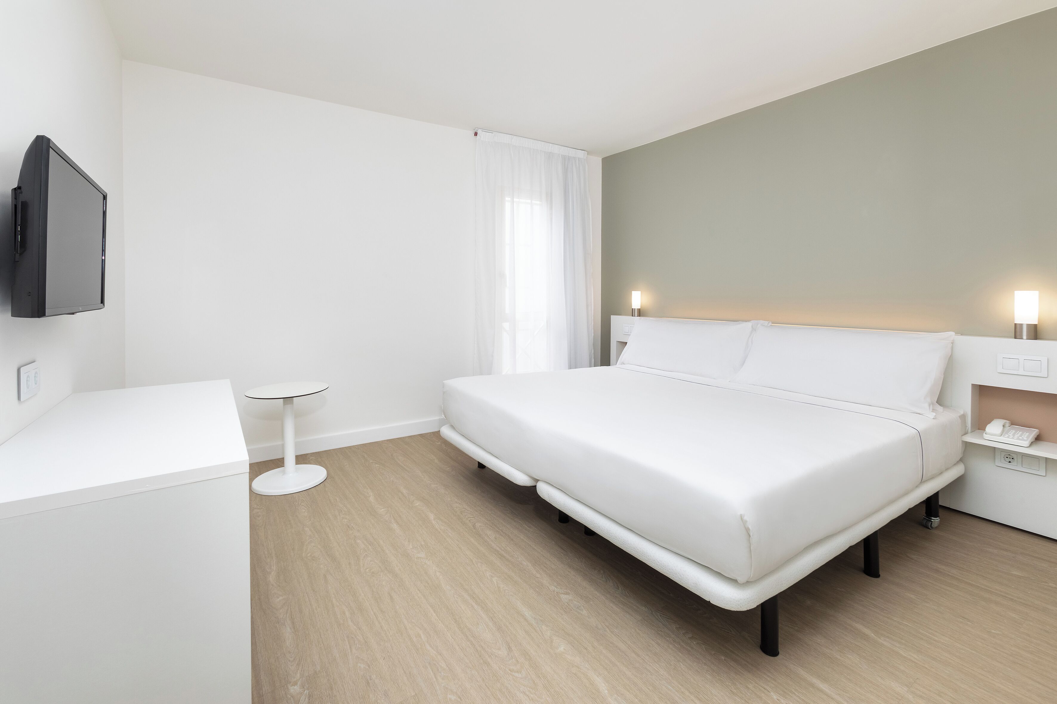 A modern hotel room with a large bed, a small bedside table, a flat-screen TV mounted on the wall, and a bedside lamp.