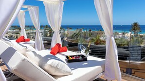 Un balcon avec des rideaux blancs, un canapé blanc et un plateau contenant des en-cas et une bouteille de champagne.