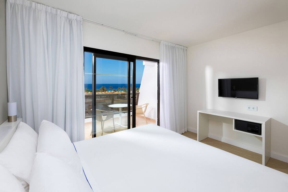 Habitación de hotel con cama, televisión, escritorio y balcón con vista al mar.