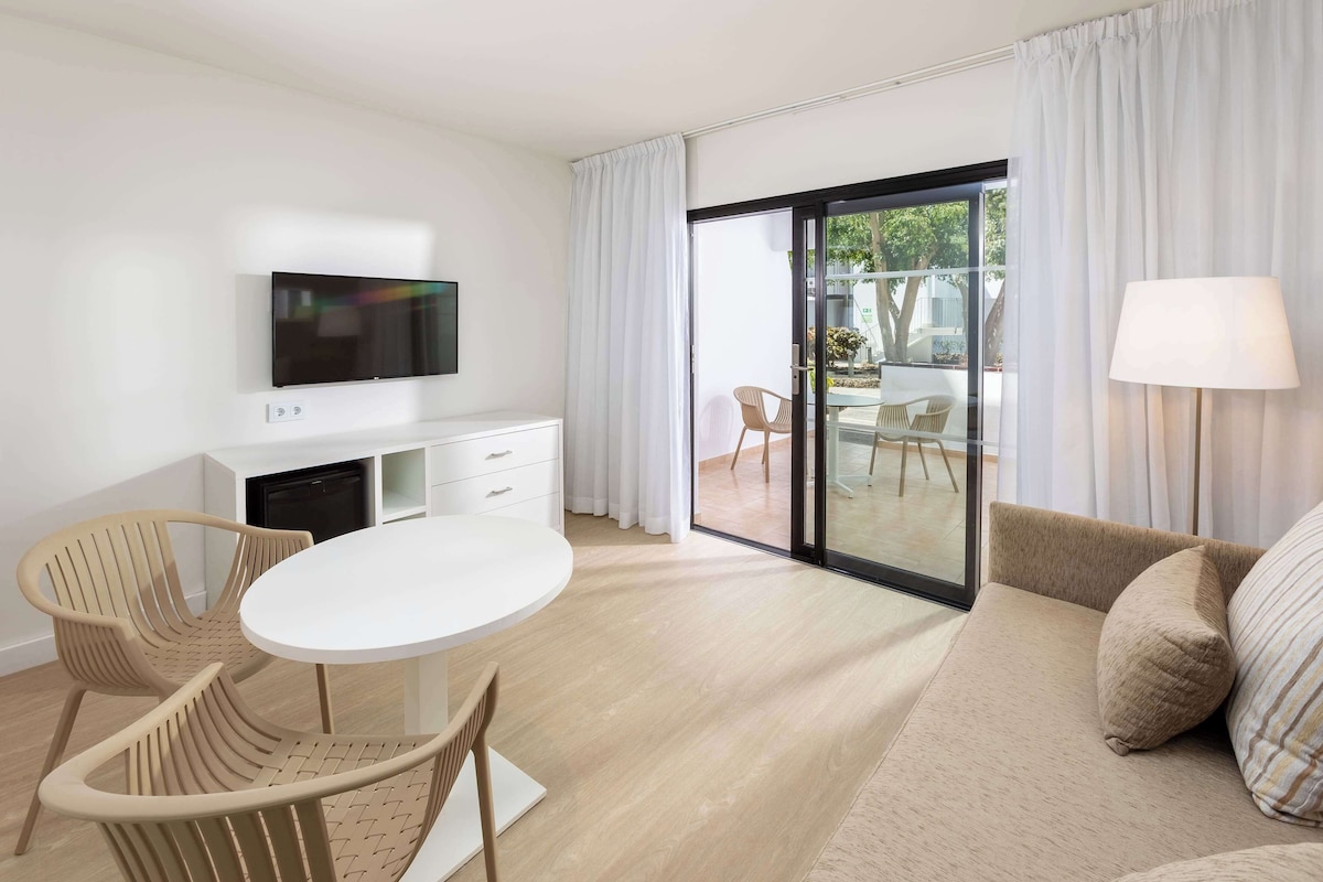 A modern living room with a sofa, a round table, and two chairs. There is a flat-screen TV mounted on the wall, a lamp, and a sliding glass door leading to an outdoor area.