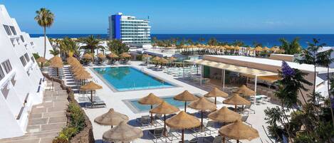 Zona de piscina d'un complex turístic amb gandules i ombrel·les, vista clara a l'oceà i un edifici gran al fons.