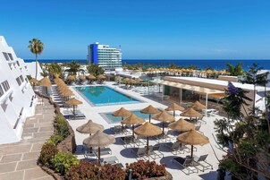 Zwembad van een resort met ligbedden en parasols, een helder uitzicht op de oceaan, en een groot gebouw op de achtergrond.