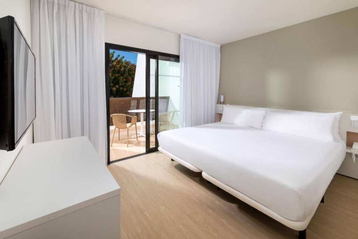 Habitación de hotel con una cama grande, televisión, balcón con mesa y sillas, y puerta de vidrio corredera.
