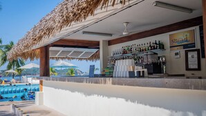 Uma área de bar com vista para a piscina, teto de palha e balcão repleto de bebidas diversas.