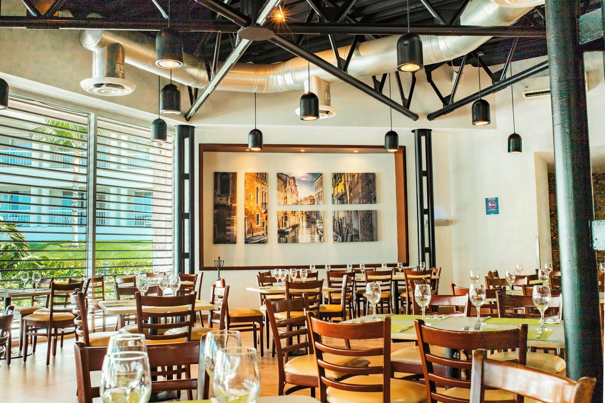 Un restaurante con mesas y sillas de madera, ventanas amplias y obras de arte en la pared.