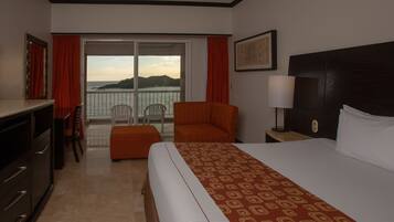 Habitación de hotel con cama, escritorio, silla, una mesita y vista al mar a través de una puerta corrediza.