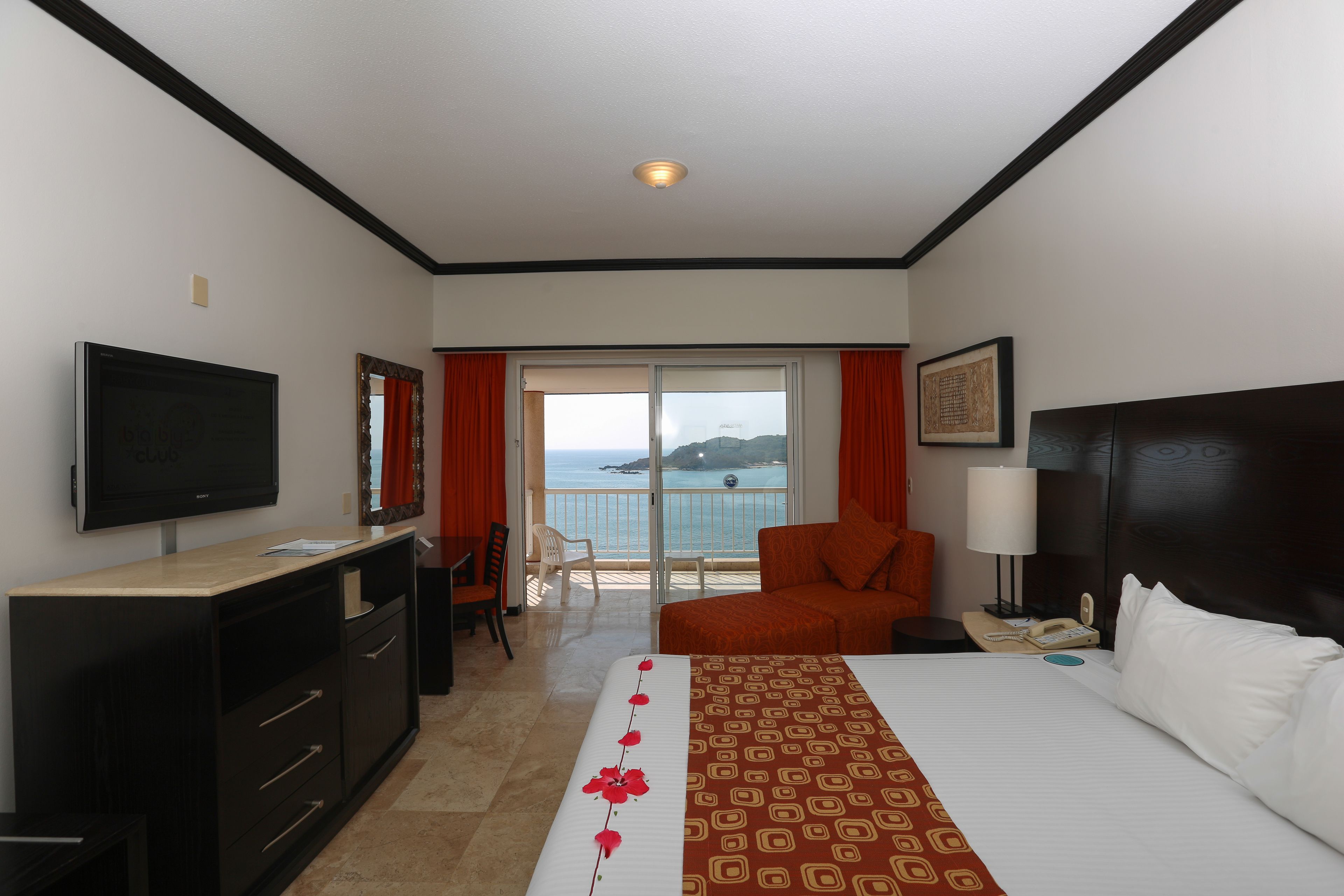 A hotel room with a bed, a desk, a television, a chair, a nightstand, and a view of the sea.
