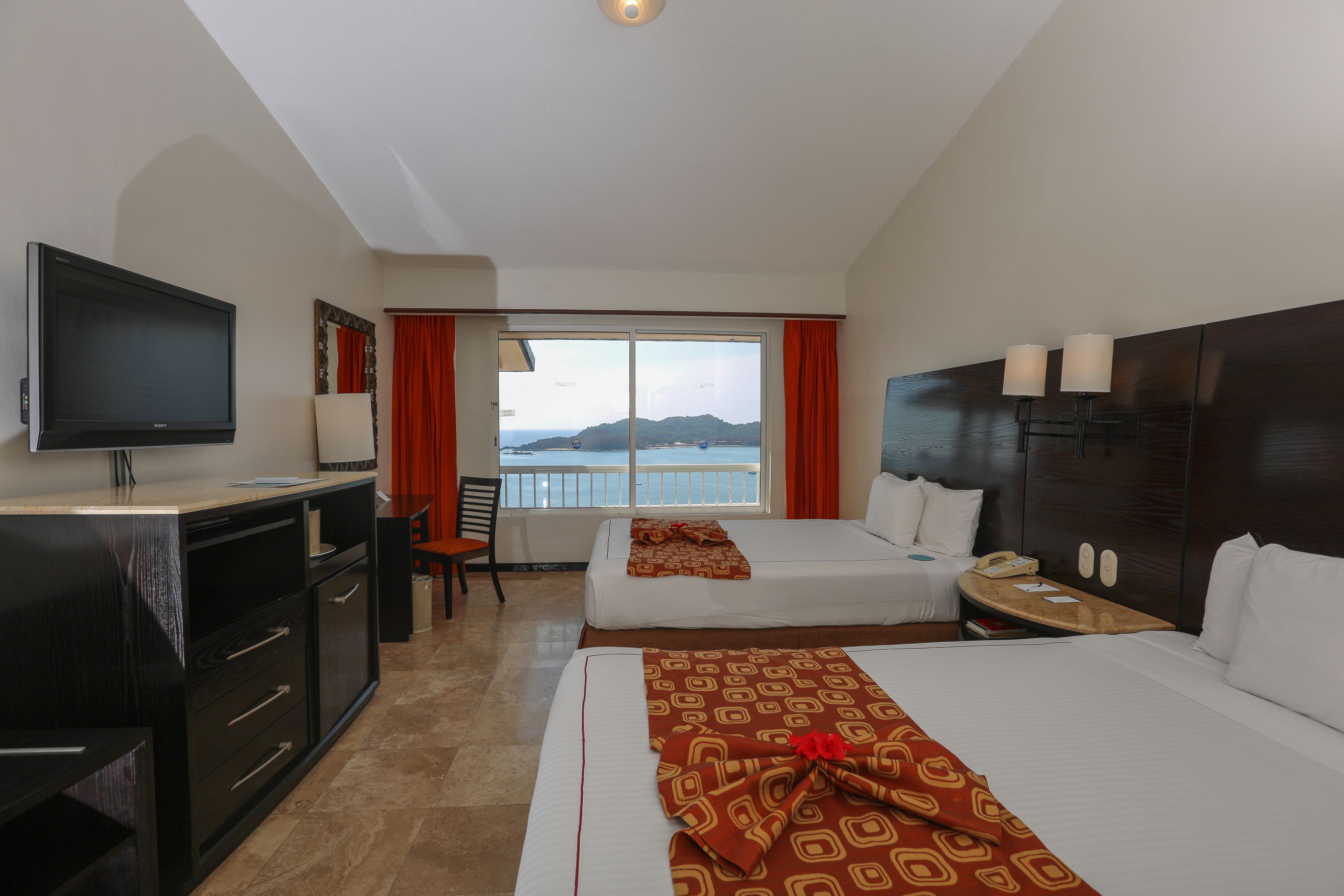 A hotel room with a TV, two beds, a desk, and a window with a view of the sea.