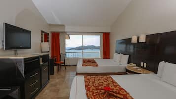 A hotel room with a TV, two beds, a desk, and a window with a view of the sea.