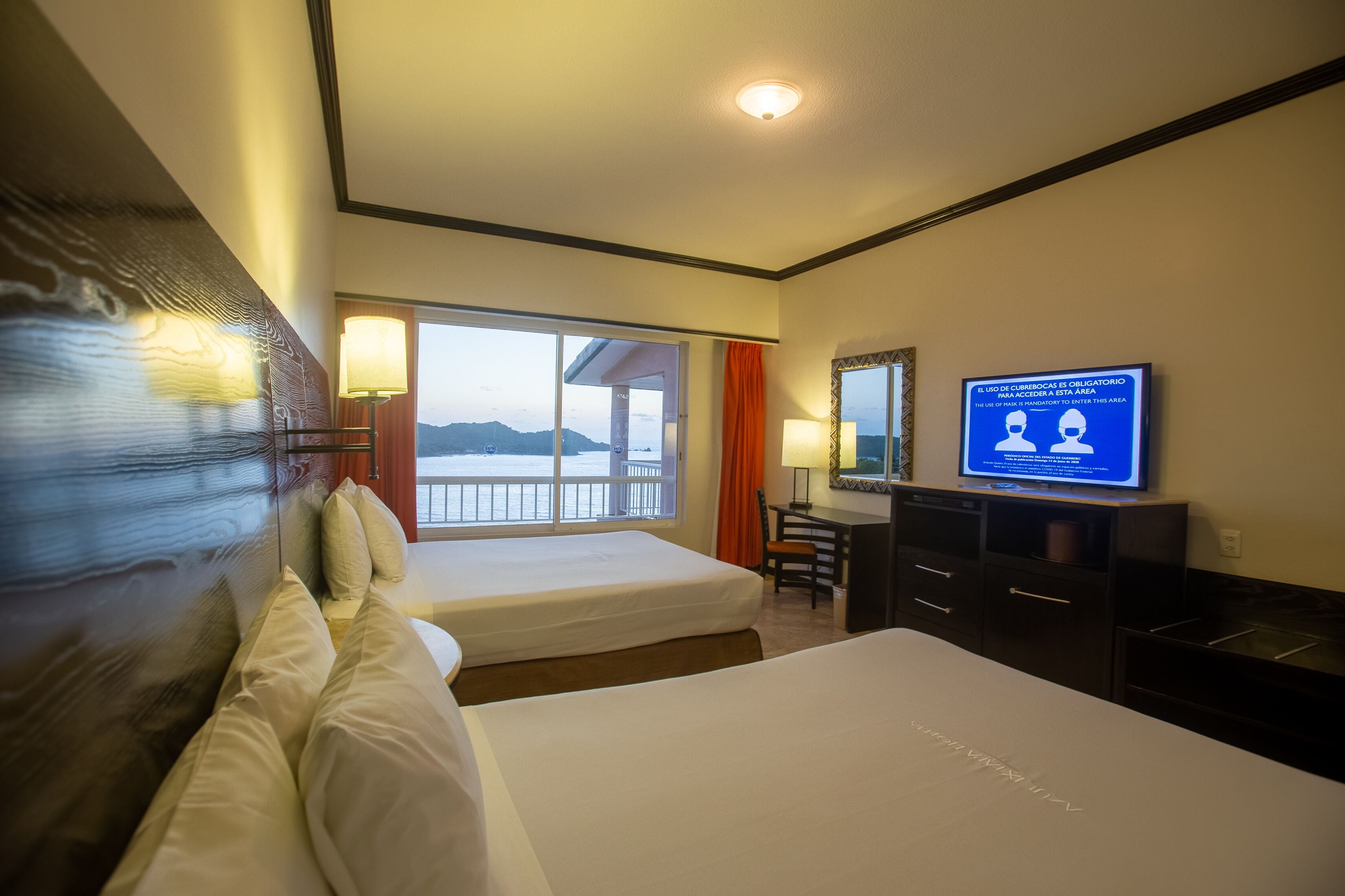 A hotel room with two beds, a desk, a television, and a large window offering a view of the mountains.
