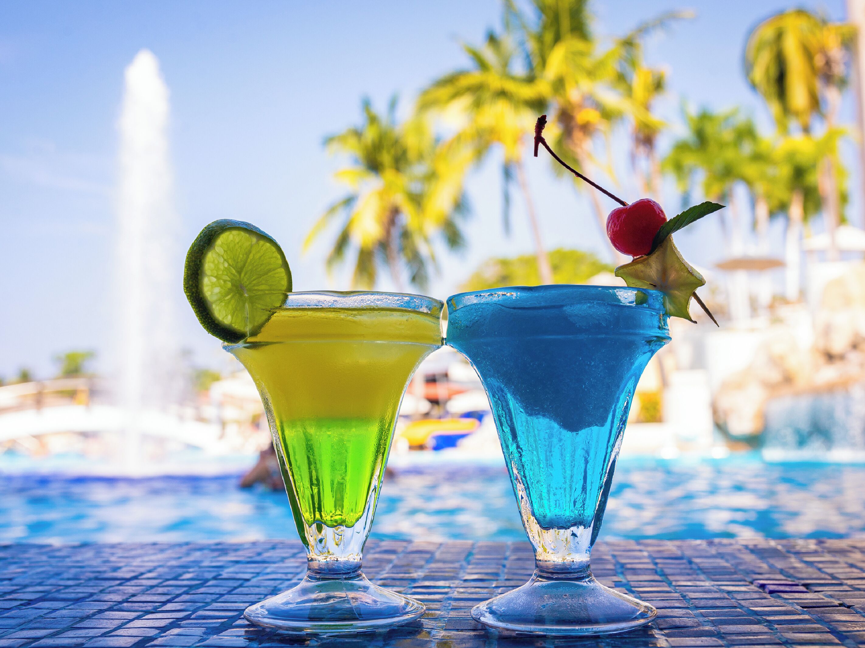 Zwei Cocktails in Gläsern, im Hintergrund ein Pool und Palmen.