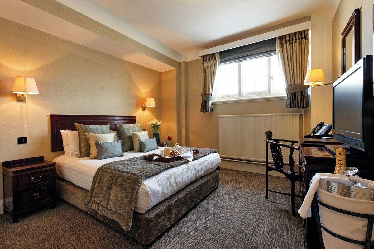 Quarto de hotel com cama, luminárias de cabeceira, escrivaninha com telefone, televisão e janela com cortinas.