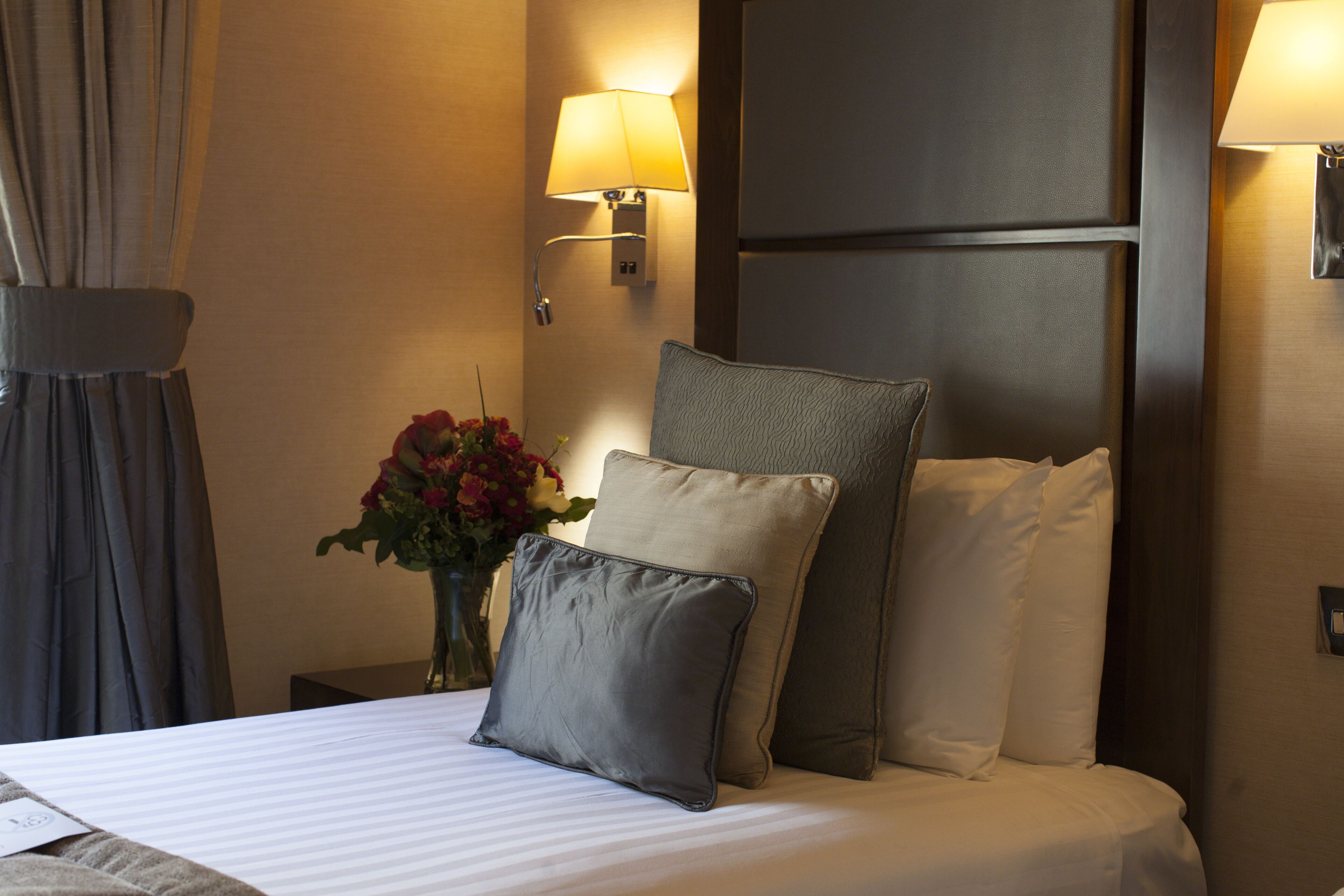 A hotel room with a neatly made bed, two wall-mounted lamps, a vase with flowers, and a curtain on the window.