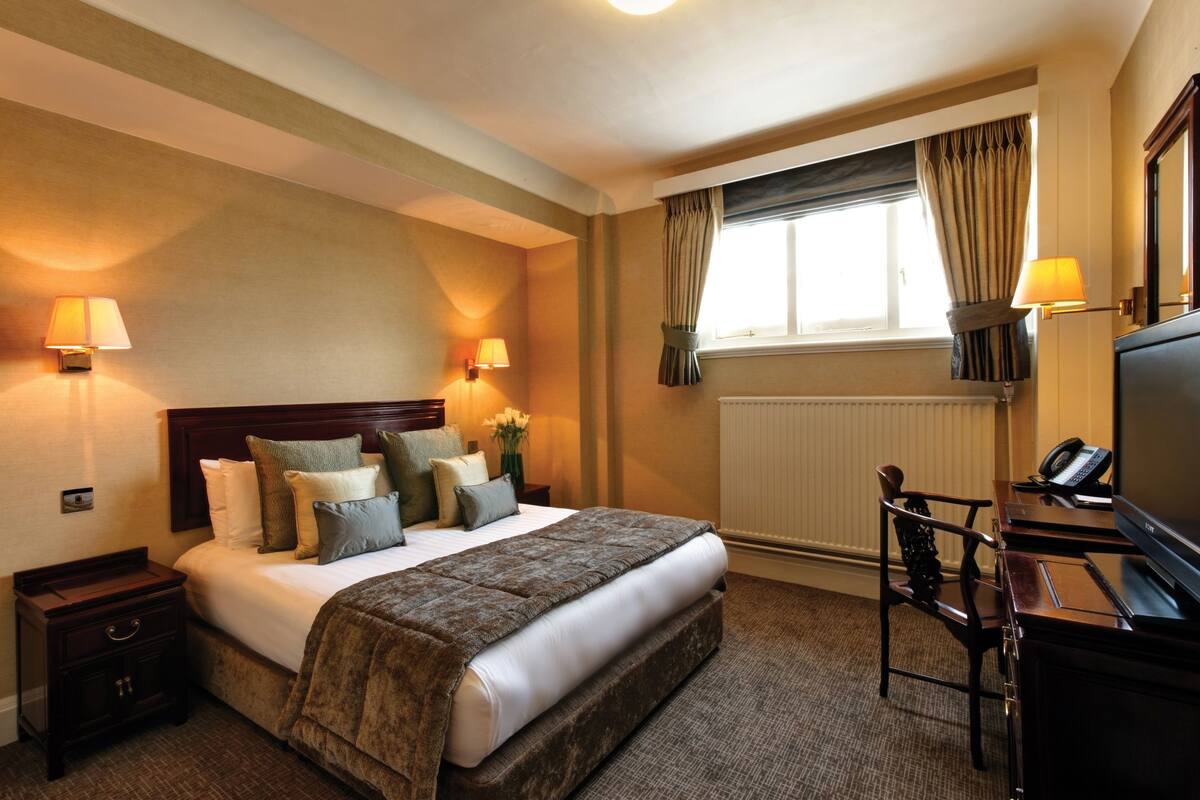Ein Hotelzimmer mit einem großen Bett, Nachttischlampen, einem Schreibtisch mit Telefon und einem fensterbepflanzten Fenster.