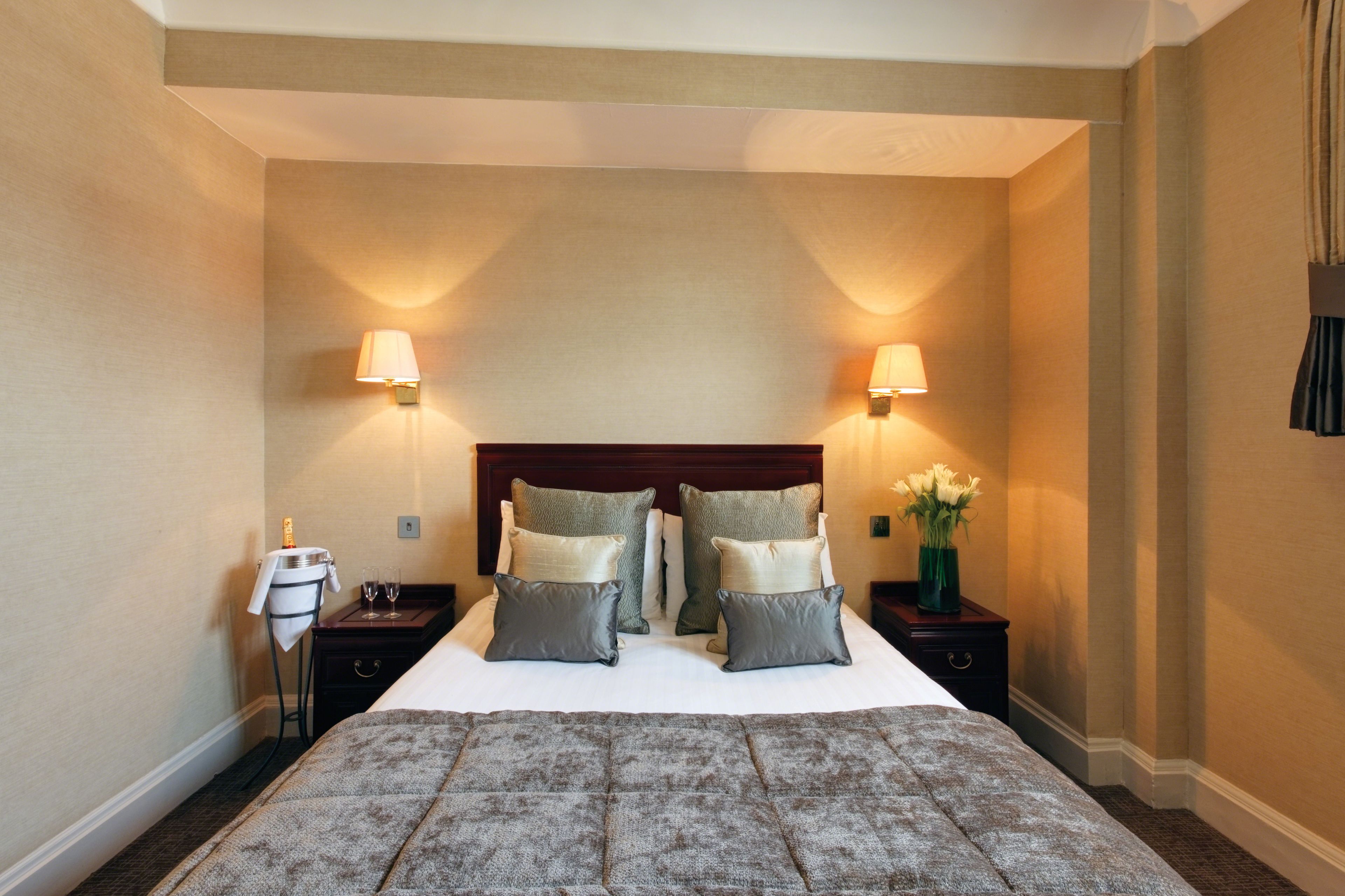 A hotel room with a large bed, two bedside tables, two wall-mounted lamps, a nightstand with a bottle and glasses, and a vase with flowers.