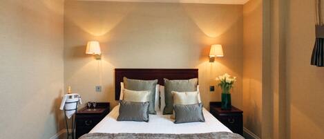 A hotel room with a large bed, two bedside tables, two wall-mounted lamps, a nightstand with a bottle and glasses, and a vase with flowers.