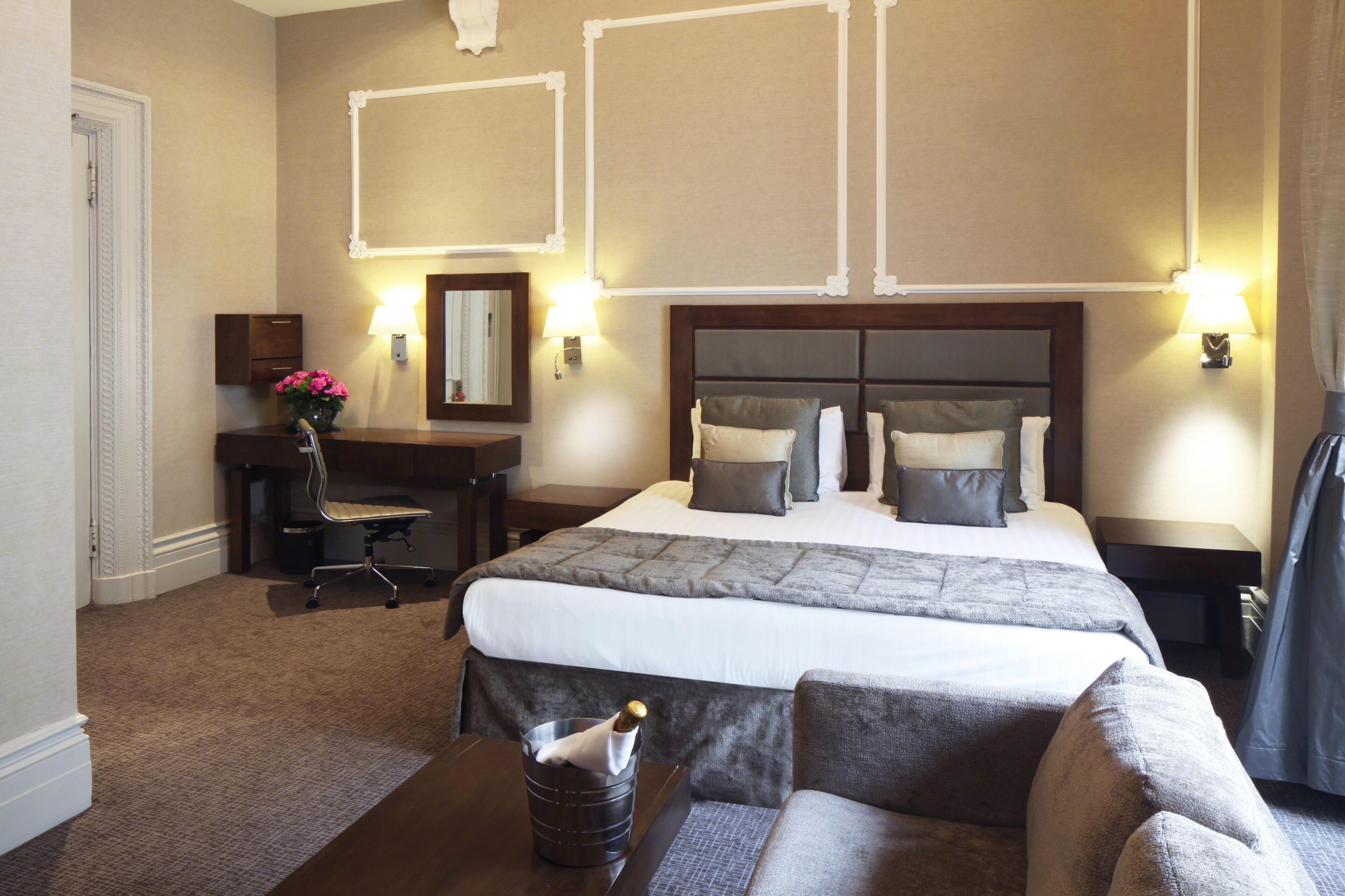 A hotel room with a large bed, a desk, a chair, and a small table with a bottle.