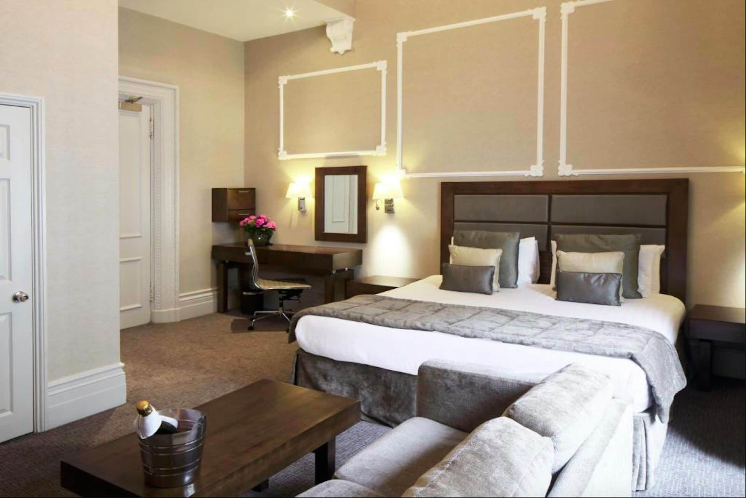 A hotel room with a large bed, a desk, a chair, and a small table with a bottle.