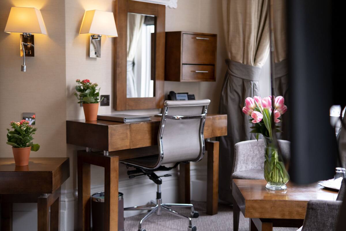 Ein Hotelzimmer mit Holzschreibtisch, einem Sessel, einem Nachttisch, einer Lampe, einem Spiegel, einer Vase mit Blumen und einem Kleiderschrank.