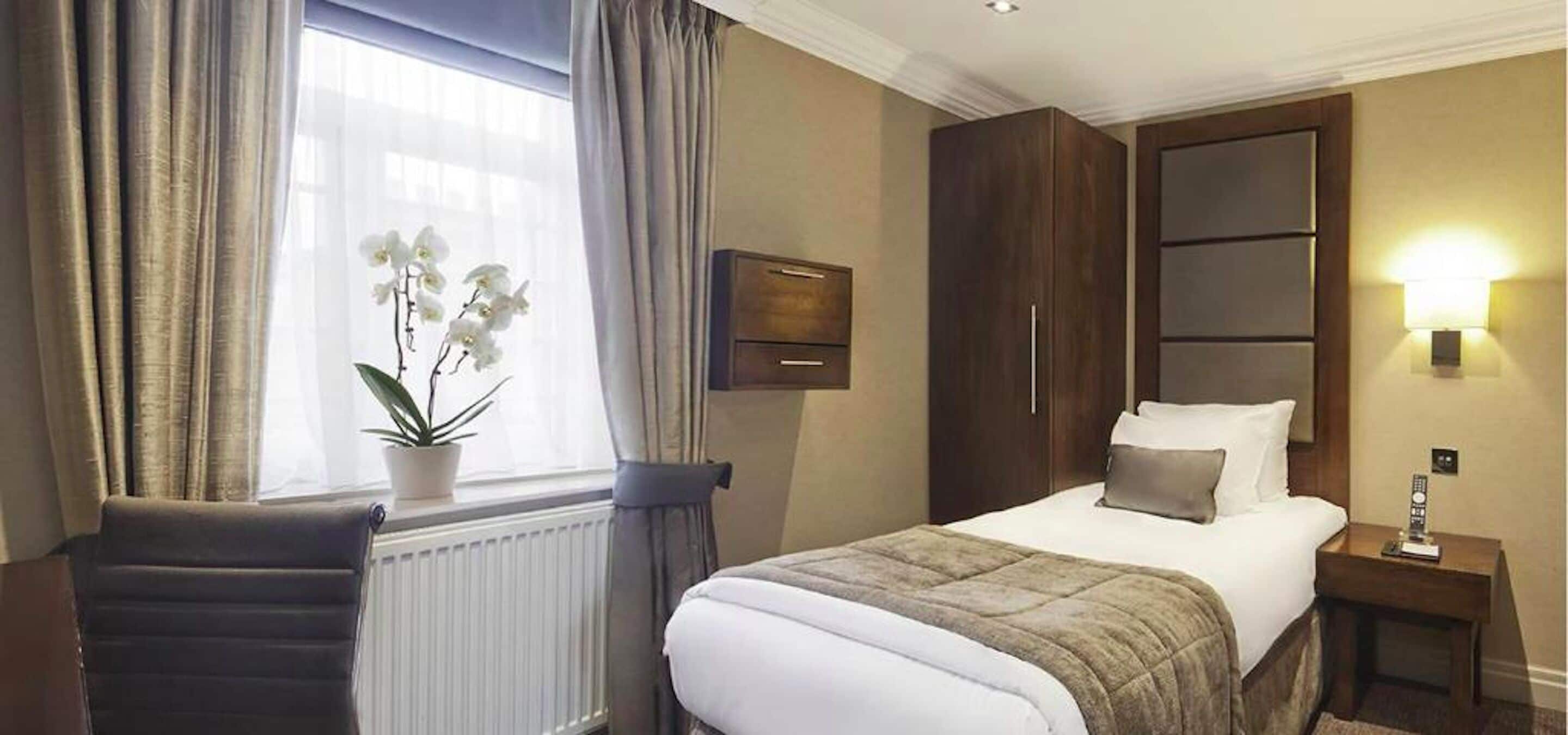 A hotel room with a bed, a nightstand, a potted plant, a chair, and a window with curtains.