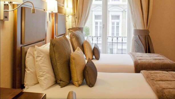 Een hotelkamer met een groot bed, meerdere kussens, een nachtkastje, een lamp en uitzicht op gebouwen door een raam met gordijnen.