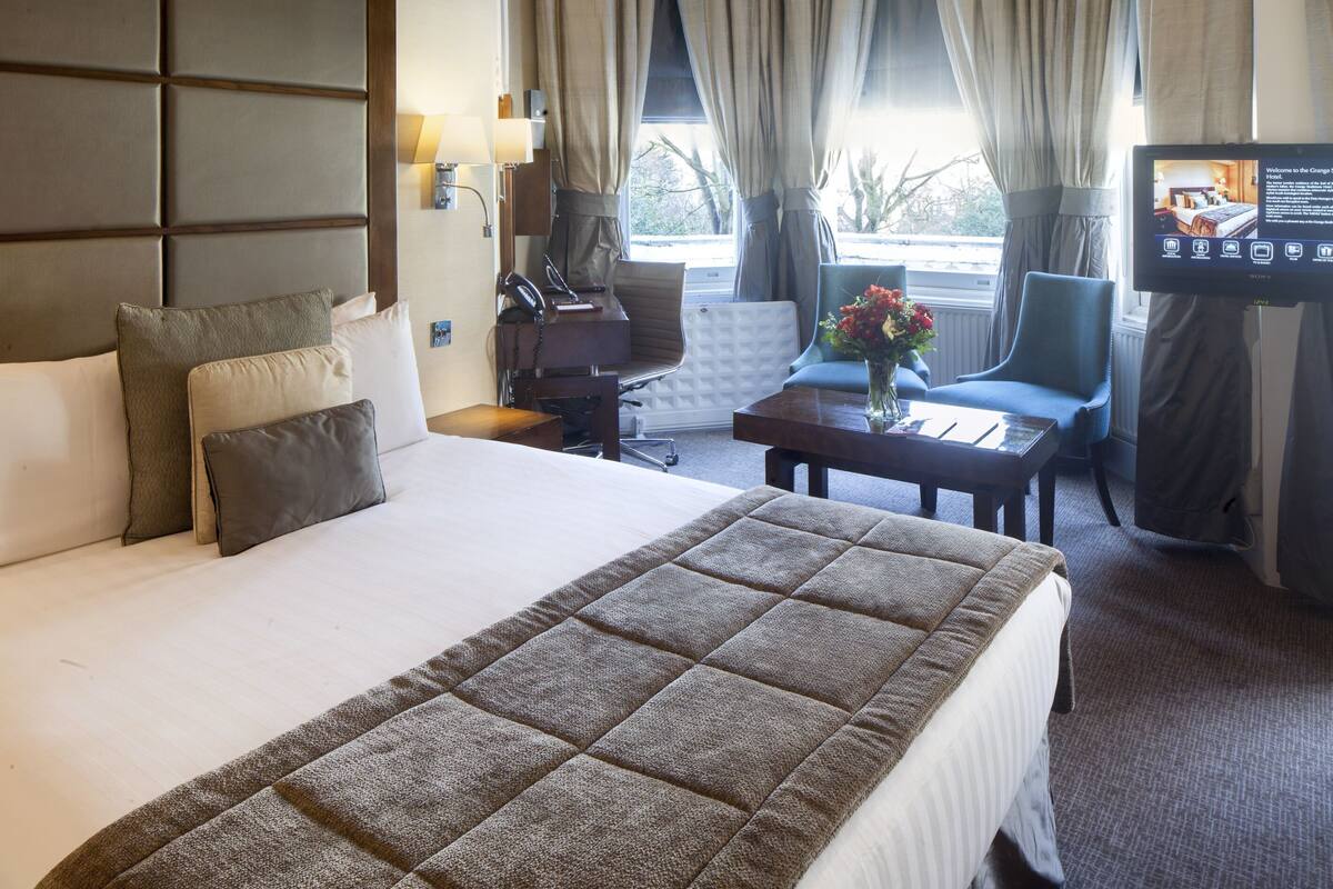 Ein Hotelzimmer mit einem großen Bett, einem Schreibtisch, einem Sessel, einem Fernseher und einem Fenster mit Vorhängen.