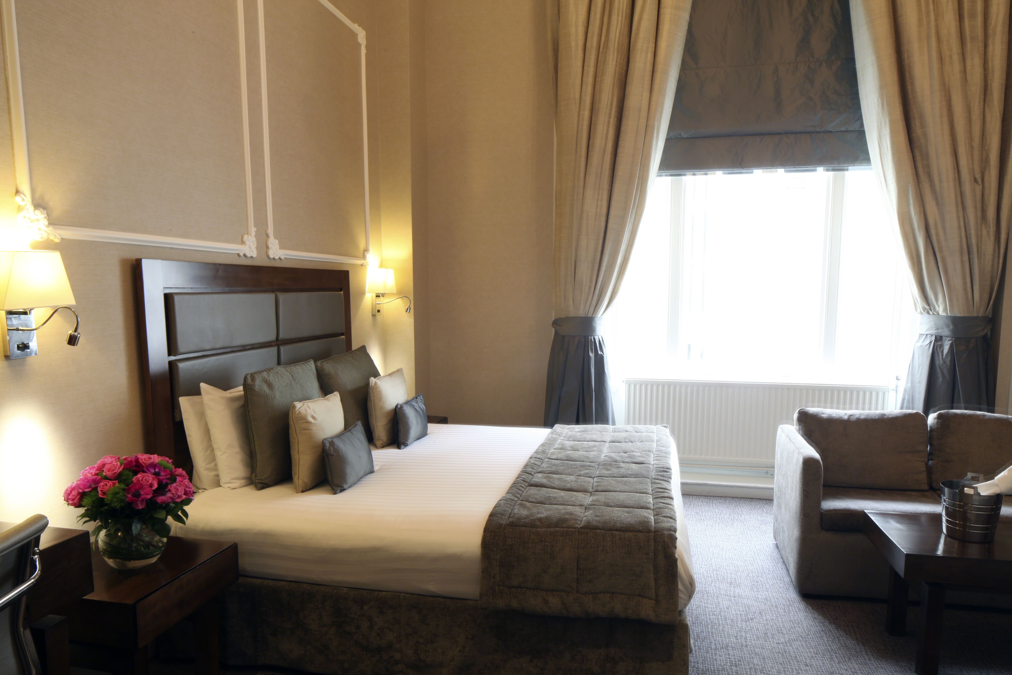 Una habitación de hotel con una cama grande, una mesita de noche con lámpara, un florero, un sofá y una ventana con cortinas.